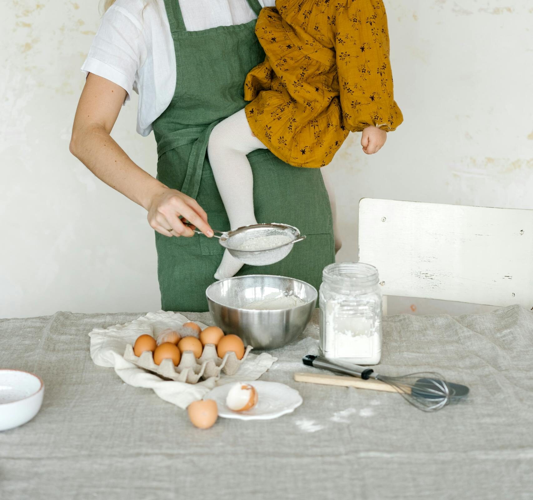 A mother and her toddler daughter baking together, adding flour to a bowl in a cozy kitchen setting.