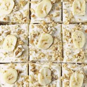 Square slices of banana bread cake with frosting and walnut garnish with banana slices.