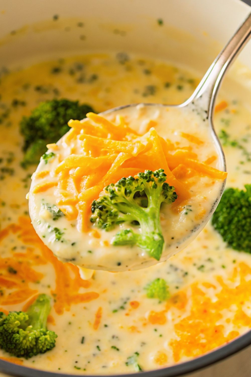A metal spoon with a spoonful of broccoli and cheese soup from a pan.